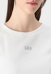 Tricou alb cu dungi, cu un decolteu larg și logo-ul "GAP" brodat în argint pe piept, având o textură netedă și un design mulat.
