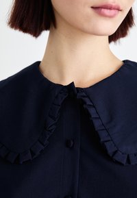 Μπλέ μπλούζα με ριγέ γιακά, κλείσιμο με κουμπιά και λεία υφή. Κοντινή λήψη που εστιάζει στις λεπτομέρειες του λαιμου και του γιακά.