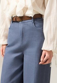 Hellblaue weit geschnittene Hosen mit einer strukturierten Oberfläche, kombiniert mit einem braunen Ledergürtel mit rechteckiger Schnalle und einer weißen Bluse mit Rüschen.
