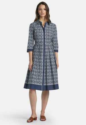 Kobieta ubrana w granatowo-białą, zapinaną na guziki sukienkę o długości do kolan z rękawami do łokci oraz brązowe sandały z odkrytymi palcami, stojąca prosto.