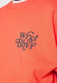 Sweatshirt arancione con una grafica testuale grigia scura "Wrst Bhvr" sul davanzale. Il tessuto sembra spesso e dalla texture liscia.