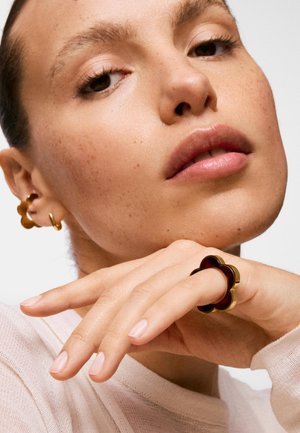 Donna con trucco naturale che indossa un anello a forma di fiore in oro e marrone scuro e orecchini coordinati in oro, poggia il mento sulla mano.