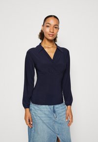 Marineblå langermet bluse med krage, kryssdesign på brystet og myk tekstur, sammen med en lyseblå jeanskjørt.