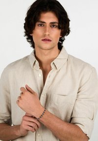 Bracciale di perle nere con accenti in argento, indossato su un polso contro una camicia di lino beige chiaro con colletto a bottoni.