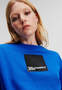 Kék pulóver, ami anyagból készült, kerek nyakkal, egy fekete négyzet alakú logóval, rajta a "KARL LAGERFELD JEANS" felirattal.