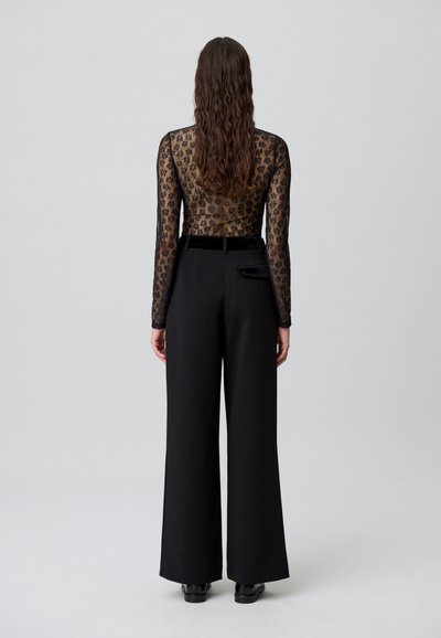 Femme aux longs cheveux bouclés, portant un pantalon large noir et un haut transparent à manches longues avec des fleurs, se tenant devant un fond uni.