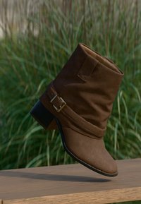 Καφέ μπότα αστραγάλου από καστόρι με διπλωμένη μανσέτα, διακοσμητική χρυσή αγκράφα και χαμηλό τακούνι τύπου block πάνω σε ξύλινη επιφάνεια, με φόντο πράσινο φύλλωμα.