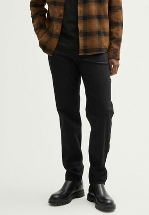 Gekacheltes braun-schwarzes Flanellhemd über einem schwarzen T-Shirt, kombiniert mit schwarzen Hosen und klobigen schwarzen Stiefeln. Fokus auf Texturen und Muster.