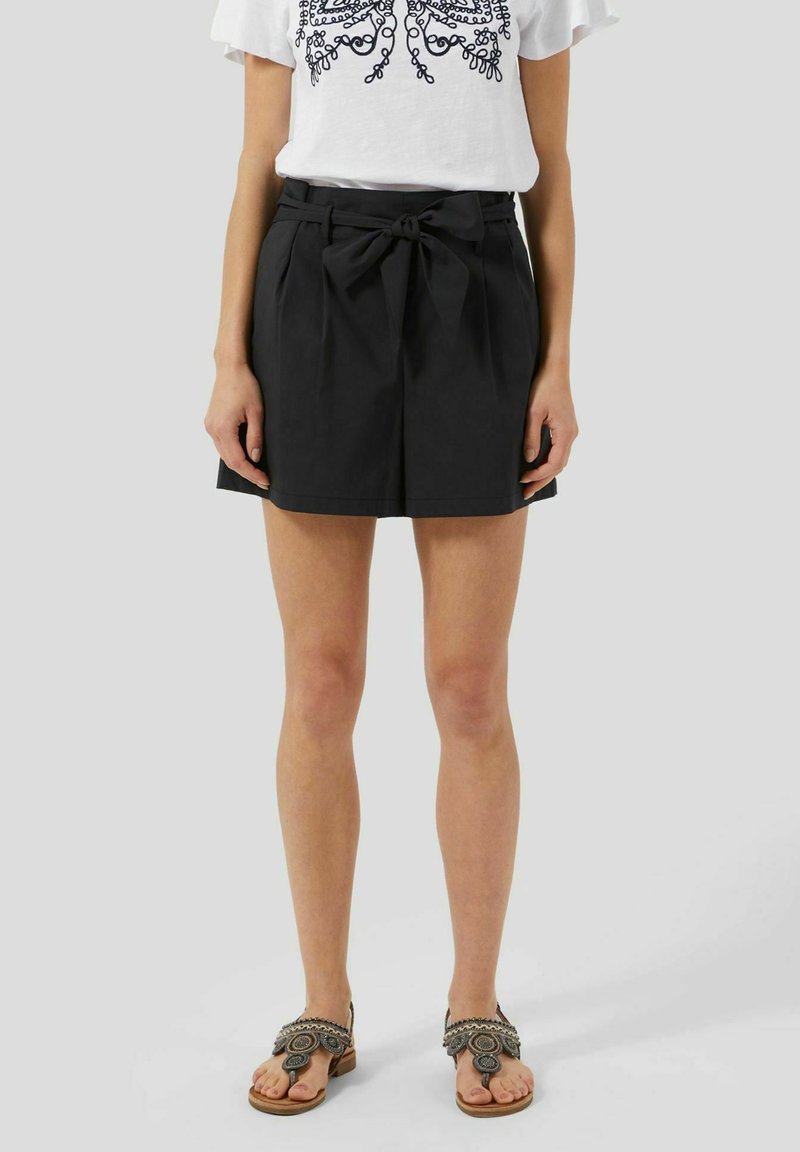 Shorts noirs taille haute avec un détail nœud à la taille, présentant des plis et une texture de tissu lisse. Associés à des sandales ornées.