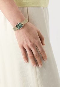 Montre rectangulaire avec un cadran vert, bracelet en maille dorée et accents en strass. Texture lisse, design moderne, reposant sur une main.
