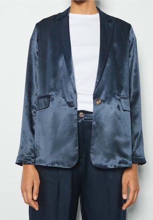 Mannequin portant un blazer en satin bleu foncé avec poches à rabat et pantalon assorti, sur un haut blanc côtelé à col rond.