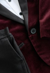 Nærbillede af en bordeaux velourblazer med sorte knapper, parret med sorte bukser og en hvid skjorte med sorte knapper.