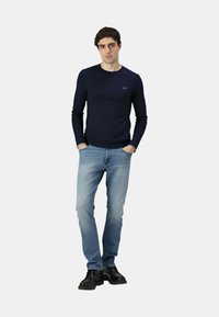 Marineblaue langärmlige Strickbluse mit taillierter Passform, kombiniert mit hellblauen Jeans und schwarzen Stiefeletten. Minimalistisches Branding auf der Bluse.