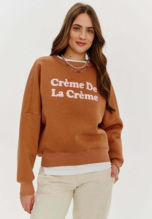Hnědá mikina s nápisem "Crème De La Crème" bílým písmem. Má pohodlný střih a žebrované manžety. Nositá přes bílé triko.