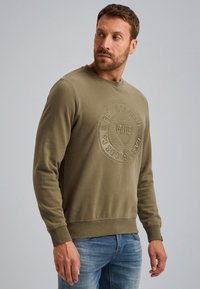 Olivengroen sweatshirt met een ronde halslijn, ribgebreide manchetten en een textured logo patch in het midden. Zacht katoen, casual fit.