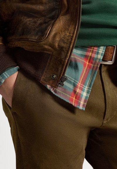 Καφέ δερμάτινο μπουφάν τύπου bomber με ριμπ μανσέτες, πράσινο πουκάμισο και καρό φανέλα σε τιρκουάζ, κόκκινο και κίτρινο. Εμφανίζονται παντελόνια ελιάς.