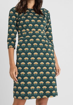 Femme portant une robe vert foncé avec un motif floral jaune et blanc répétitif et des manches trois-quarts.