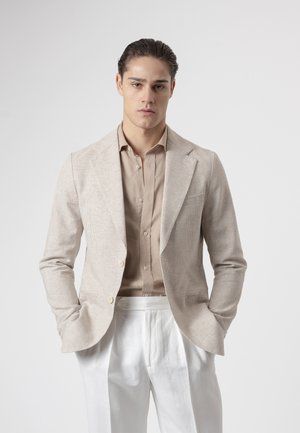 Joven con cabello peinado hacia atrás que lleva un blazer beige texturizado, camisa marrón claro y pantalones blancos, con las manos en los bolsillos y expresión neutral.