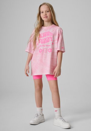Młoda dziewczyna z długimi włosami, nosząca za dużą różową koszulkę w stylu tie-dye z napisem "Always Sunny", różowe szorty, białe skarpetki i białe trampki.