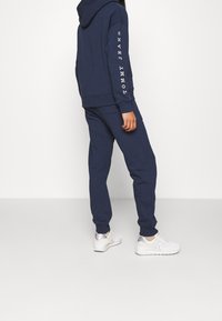 Mörkblå hoodie och sweatpants-set i mjukt tyg, med en logotyp på ärmen, ihop med vita träningsskor.