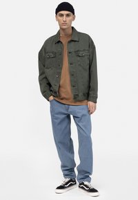 Olive green denim jacket with flap pockets, brown crewneck shirt, light blue straight-leg jeans, and black sneakers with white soles.
