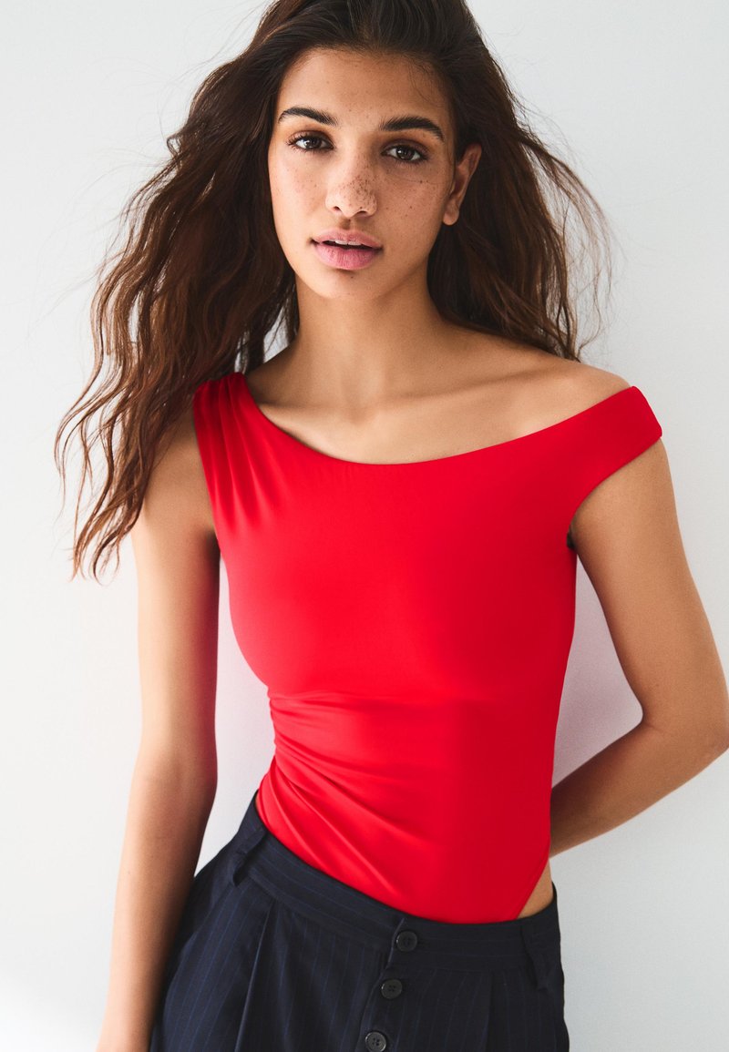 Jeune femme aux longs cheveux ondulés portant un haut rouge décolleté aux épaules et un pantalon sombre taille haute, debout devant un fond blanc uni.