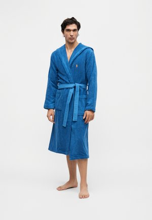 Homme debout pieds nus portant un peignoir à capuche bleu noué à la taille avec deux poches avant sur un fond clair uni.