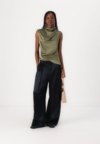 Un haut sans manches, drapé, de couleur vert olive, associé à un pantalon large bleu marine. Modèle tenant un sac à main beige avec un détail de fermoir.