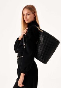 Borsa tote in pelle nera embossata con una forma strutturata, parte superiore curva e dettagli in metallo dorato, dotata di doppie maniglie e motivi testurizzati.