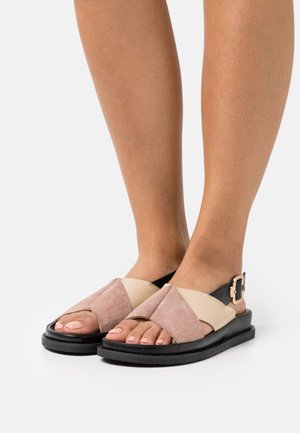 Des pieds portant des sandales en suède beige et rose à lanières croisées avec des semelles noires et une sangle noire sur un fond blanc.