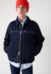 Sötétkék farmerdzseki sárga varrással, cipzárral záródik, és mellzsebe van. Világoskék galléros ing felett viselik. Élénk piros beanie sapka.