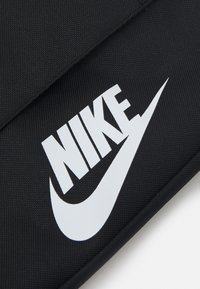 Melna auduma tekstūra ar izteiktu virsmu, kurā ir izcils balts Nike logo un vēlme. Logo ir drosmīgs un labi definēts.