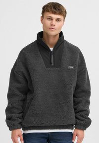 Fleece pullover in dark gray with a half-zip front, featuring a smooth black panel and a small logo on the chest. Soft texture and relaxed fit.