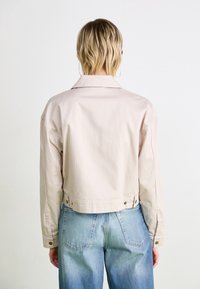 Cropped beige jacket with subtle pinstripes, long sleeves, and a classic collar. Features snap buttons at the waist and cuffs, paired with blue jeans.