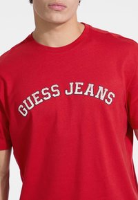 Raudona medvilninė trumpomis rankovėmis marškinėliai, su apvaliu kaklo iškirpimu ir baltu nusidėvėjusiu užrašu "GUESS JEANS" per krūtinę.