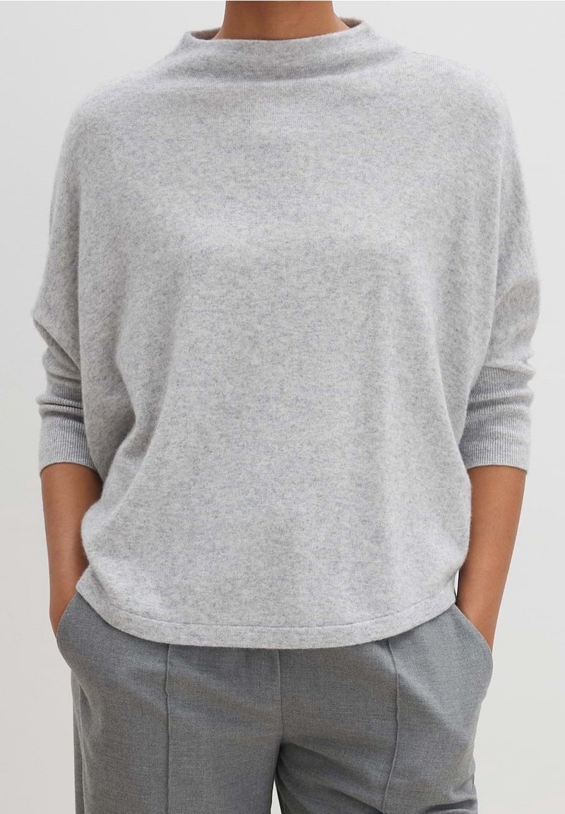 Personne portant un pull en maille gris clair à manches longues, coupe ample et col montant, associé à un pantalon gris sur mesure avec les mains dans les poches.