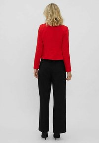 Veste rouge courte avec manches longues, texture lisse et coupe ajustée, associée à un pantalon noir à jambes larges et des chaussures à talons hauts.