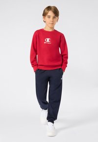 Rode sweatshirt met wit Champion-logo, gecombineerd met marineblauwe sweatpants en witte sneakers. Glad textuur, casual pasvorm, ribgebreide boorden.