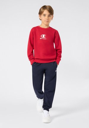 Rotes Sweatshirt mit weißem Champion-Logo, kombiniert mit dunkelblauen Sweatpants und weißen Sneakers. Glatter Stoff, lässige Passform, gerippte Bündchen.
