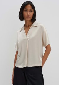 Helle beige Bluse mit kurzen  Ärmeln und Kragen, mit einem V-Ausschnitt-Design, glatter Textur und lockerer Passform. Kombiniert mit schwarzen Hosen.