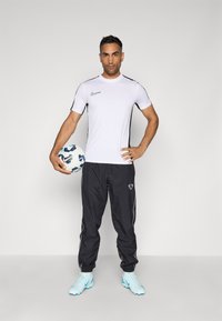Weißes kurzärmeliges Sportshirt mit schwarzen Akzenten, schwarze Jogginghosen und hellblaue Fußballschuhe, hält einen gemusterten Fußball.