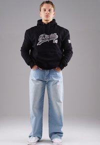 Schwarzer Kapuzenpullover mit weißem "Picaldi 23"-Text, kombiniert mit hellblauen weit geschnittenen Jeans. Das Model trägt weiße Sneaker. Einfacher Hintergrund.
