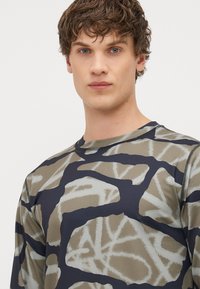 Camisola desportiva de manga longa em padrão camuflado, apresentando tons de verde e azul-marinho. Feita de um tecido liso e elástico com gola redonda.