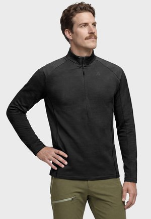 Mann mit Schnurrbart trägt eine schwarze, strukturierte Jacke mit Reißverschluss und olivgrüne Hose und steht mit einer Hand in der Hüfte vor einem einfarbigen Hintergrund.