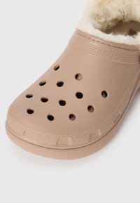 Beige tresko laget av glatt plast, med ventilasjonshull, en teksturert såle og en fluffy innvendig fôr.