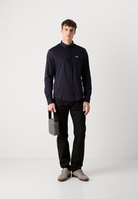 Marineblå langermet skjorte med krage og knapper, slim fit. Sorte bukser og grå veske med håndtak. Grå joggesko med snøring og subtile detaljer.