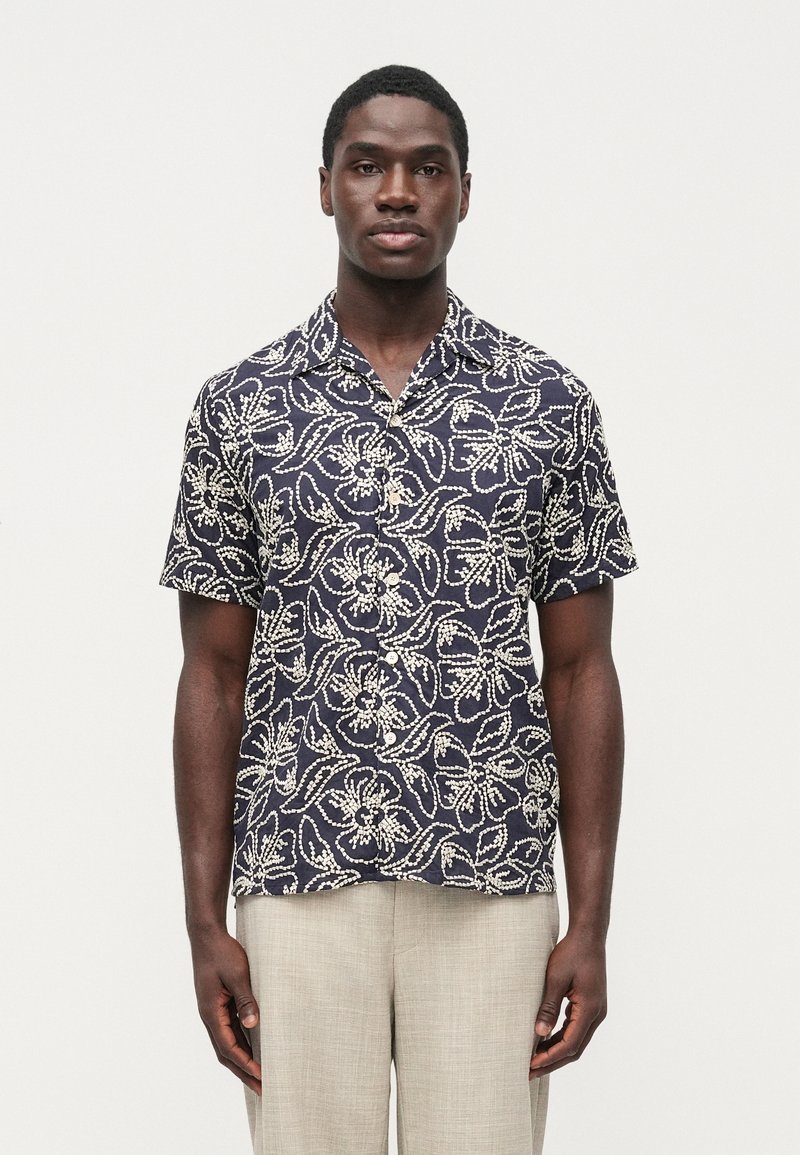 Košeľa s krátkym rukávom z námorníckej modrej látky s bielymi kvetinovými a listovými vzormi. Zapínanie na gombíky vpredu, voľnejší strih a golier.