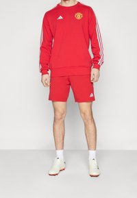 Persona che indossa una maglietta a maniche lunghe rossa del Manchester United e pantaloni abbinati, con scarpe e calze bianche Adidas su uno sfondo neutro.