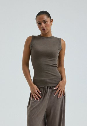 Žena s vlasmi v cope, oblečená v hnedom bezrukávovom rebrovanom topán a vo voľných hnedých nohaviciach so šnúrkou, stojaca s rukami opretými o boky.