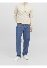 Ljusbeige dragkedjesweatshirt med svart text, lager-på-lager över en brun skjorta, matchad med lösa blå jeans och vita sneakers.